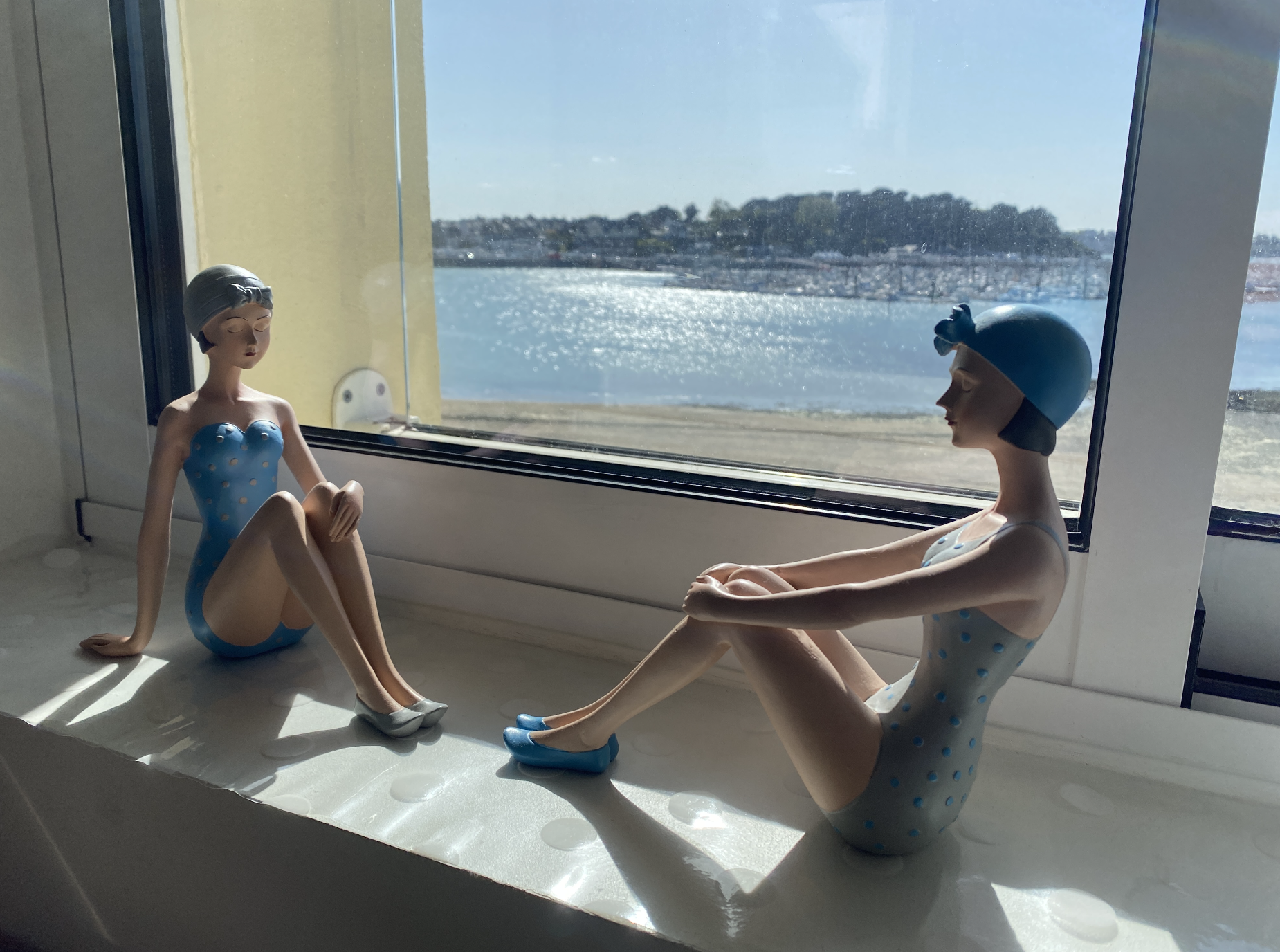 Lumière du soleil et lumière du ciel entrent à travers une fenêtre avec vue sur mer. Elle sont symbolisées par deux statuettes de baigneuses